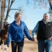 John and Leslie Malone take a walk on their property, holding hands, with a horse and female rider in the background.