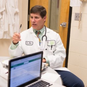 Dr. Craig Webb speaks to students while sitting in a chair in his scrubs veterinary coat