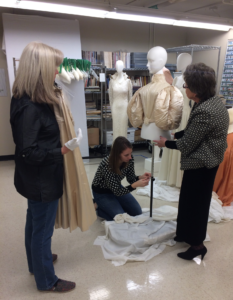 Volunteers Jan Alexander (left) and Mary Biggers (right) assist acting curator/collections manager Megan Osborne in dressing a mannequin with the 1890 wedding gown, following Mary's careful work to reattach the original leg-of-mutton sleeves to the bodice.