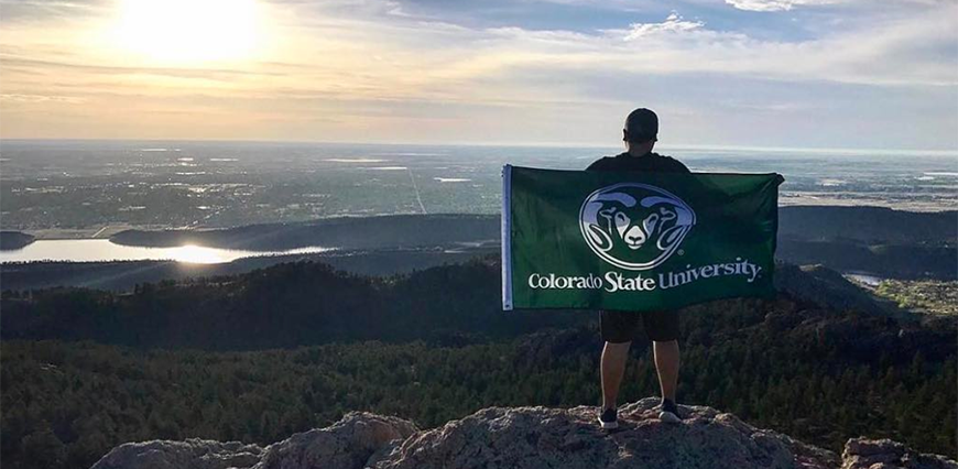 Guy standing on top of Horsetooth Rock in Fort Collins holding out a CSU flag