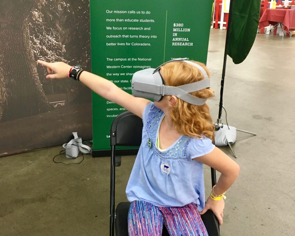 Young girl points into the distance wearing virtual reality headset at CSU's booth at the 2018 Denver County Fair.