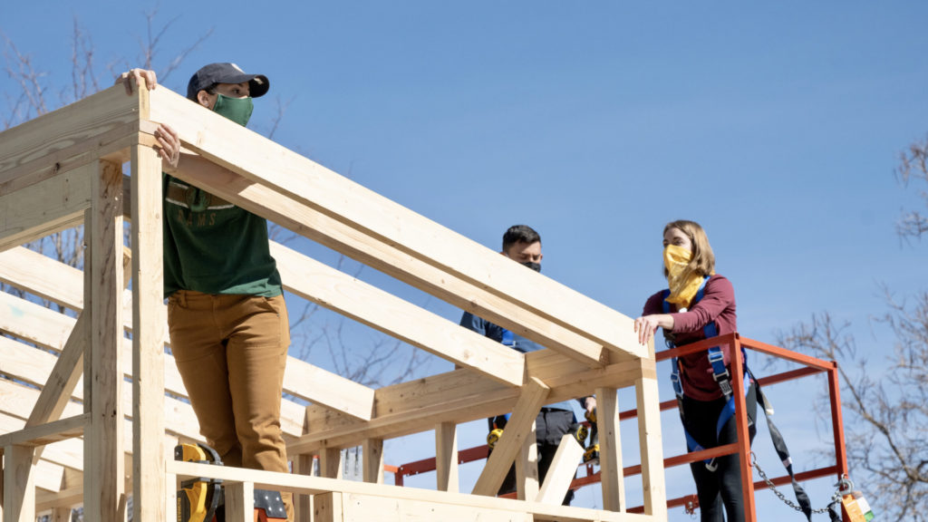 Students stand on scaffolding and wooden frame of tiny house on wheels, during construction on sunny day at the Nancy Richardson Design Center, March 6, 2021.