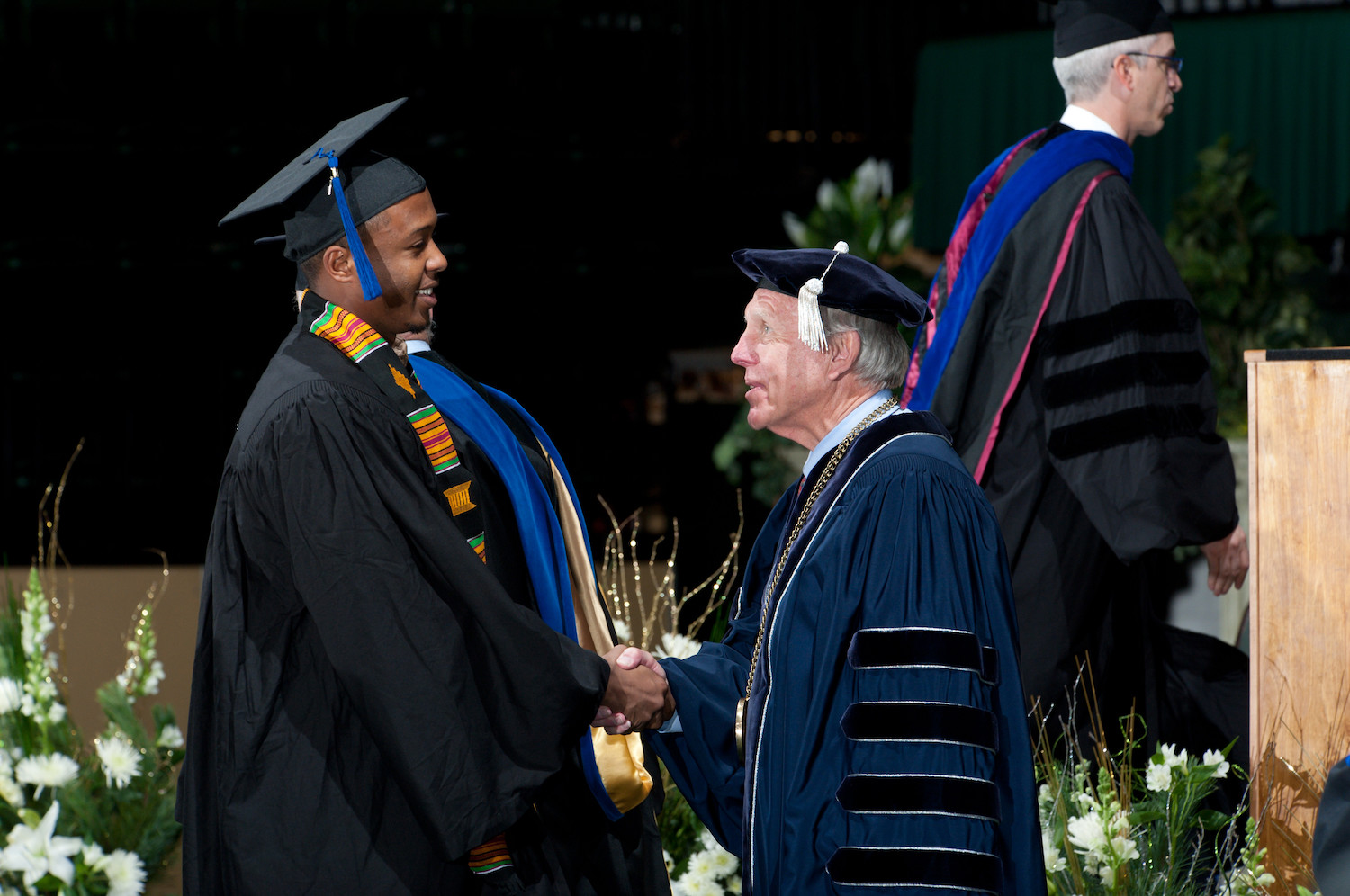 Man in academic regalia shakes the hand of a graduating student in a cap and gown.