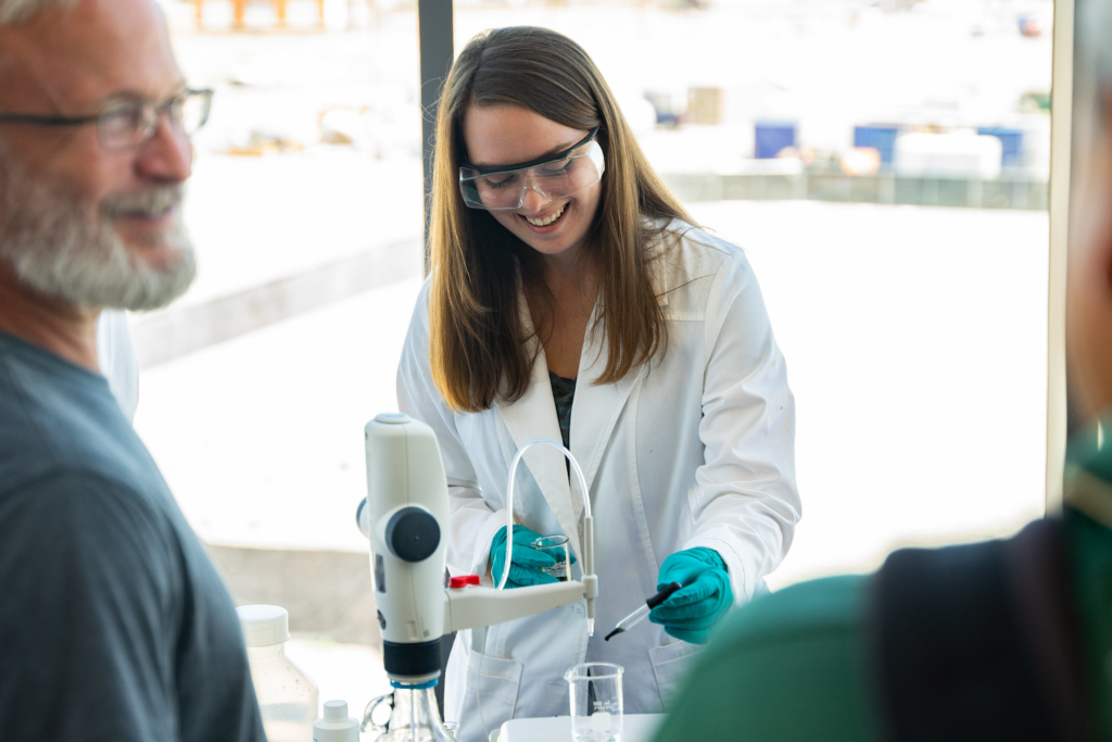 A young woman in a lab coat uses a dropped to add liquid to a beaker.
