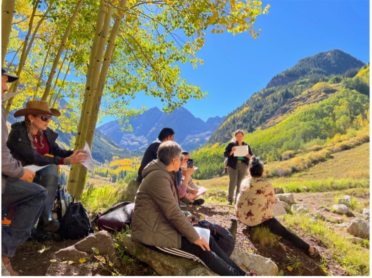 A group of six people listen to a lecture with mountains in the background.