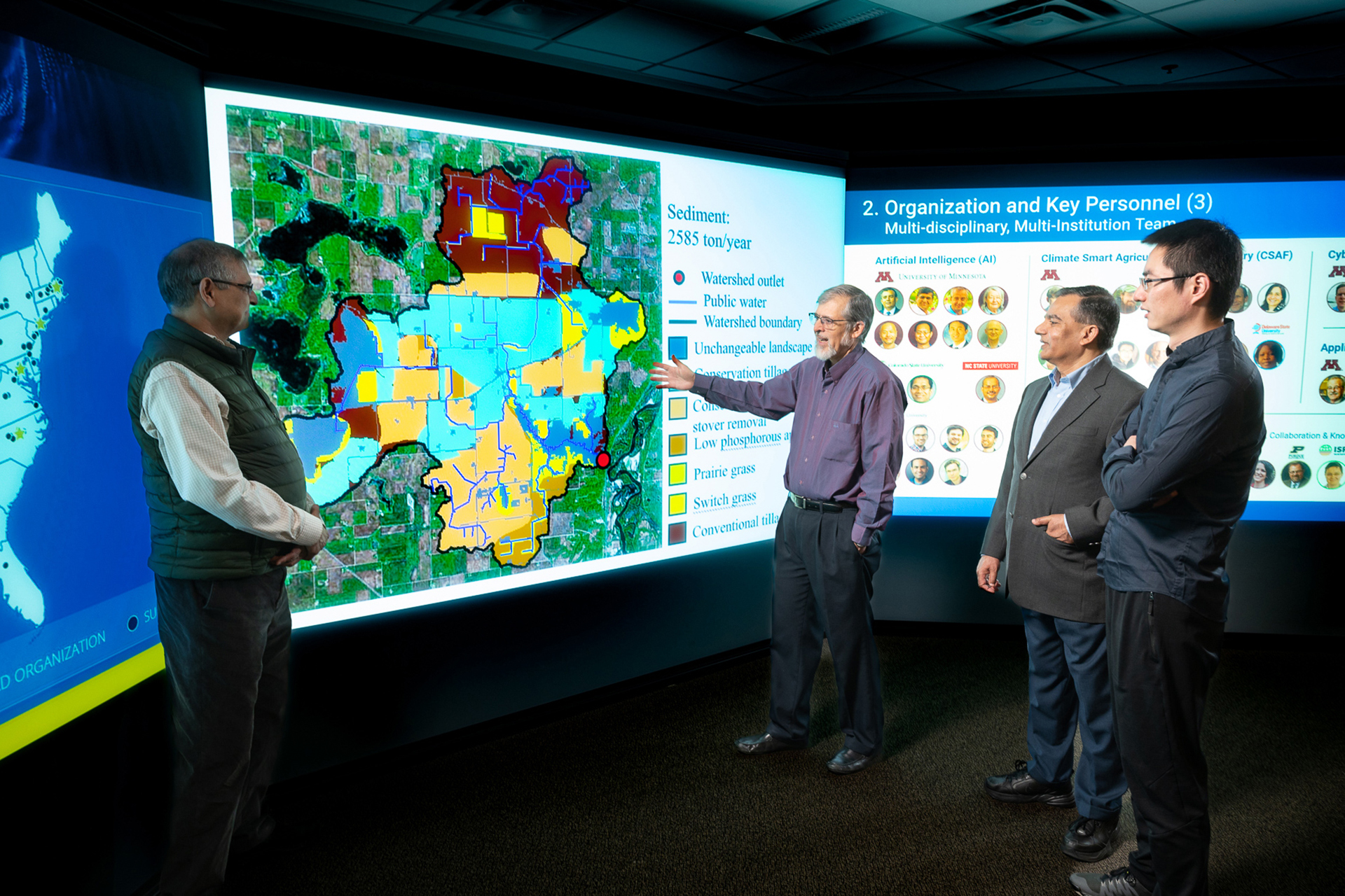 Four men stand before a projected map and one explains research that is displayed on the screen