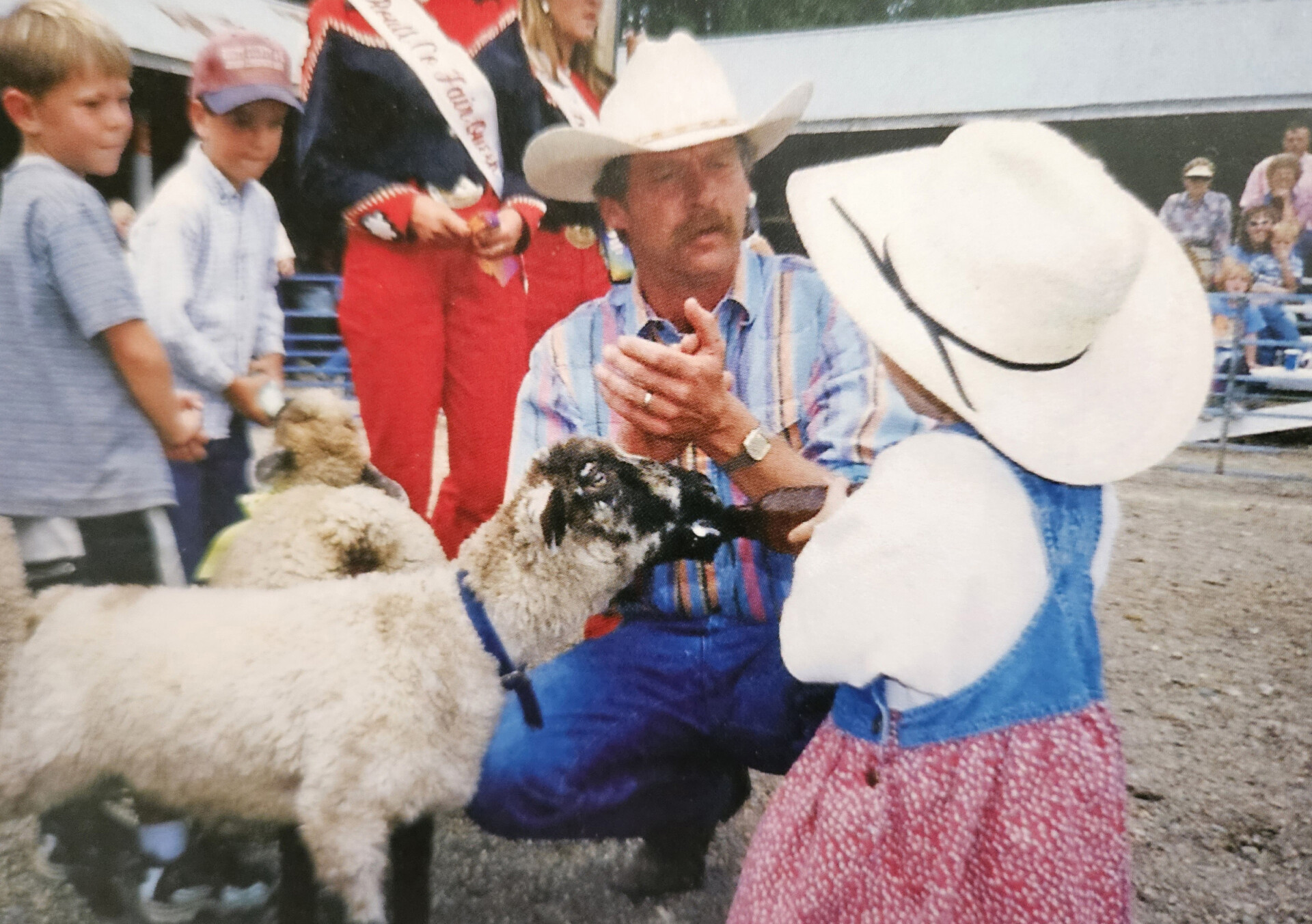 A man in a cowboy hat kneels down and talks to a young girl bottle-feeding a lamb