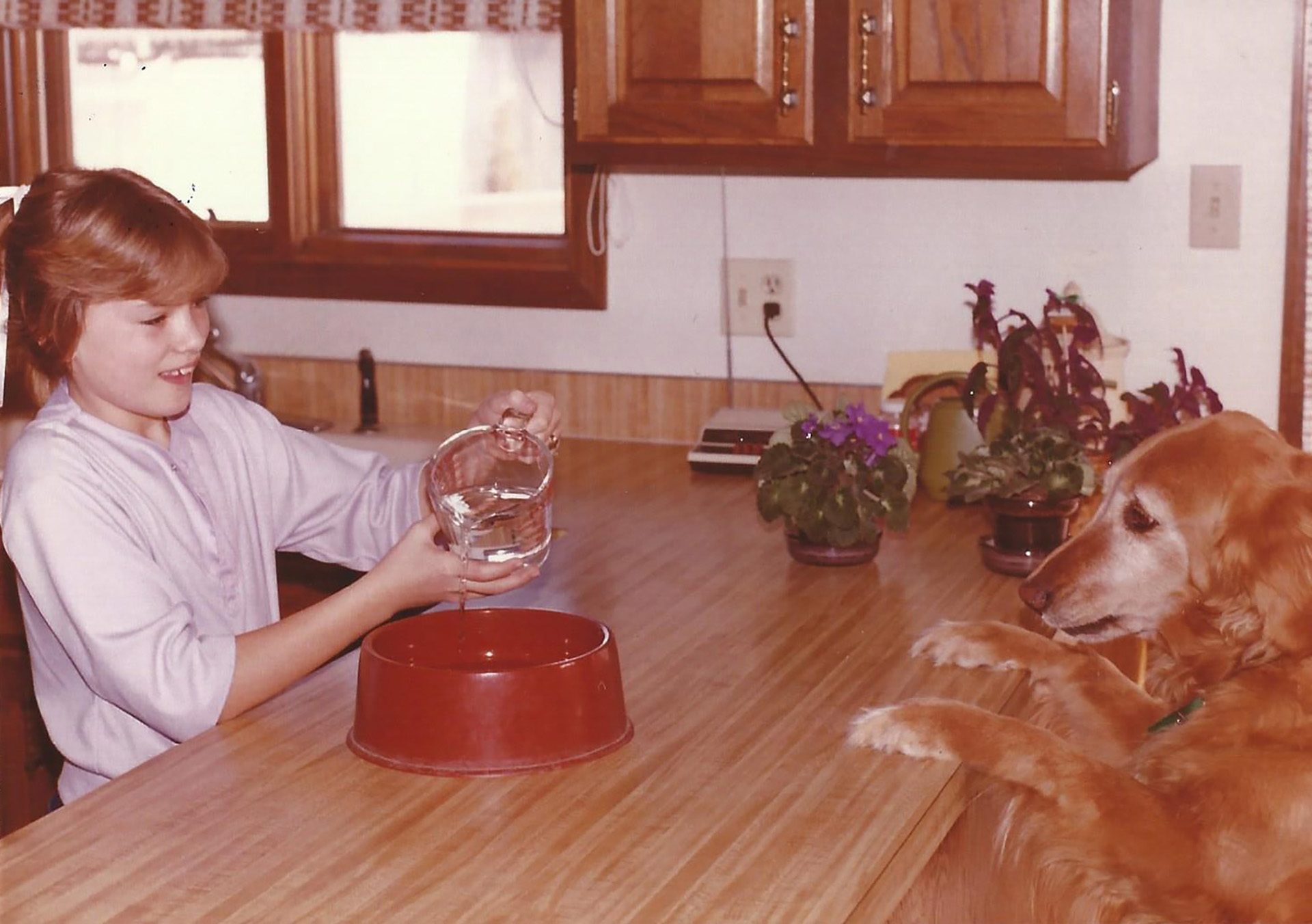 A girl pours water into a dog bowl sitting on a kitchen counter as her dog watches