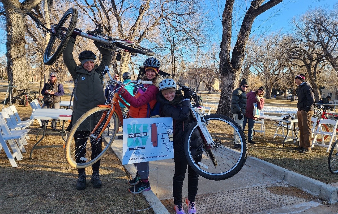 Three young women hold their bicycles in the air near a sign saying Bike to Work Day, dressed for cold weather outdoors