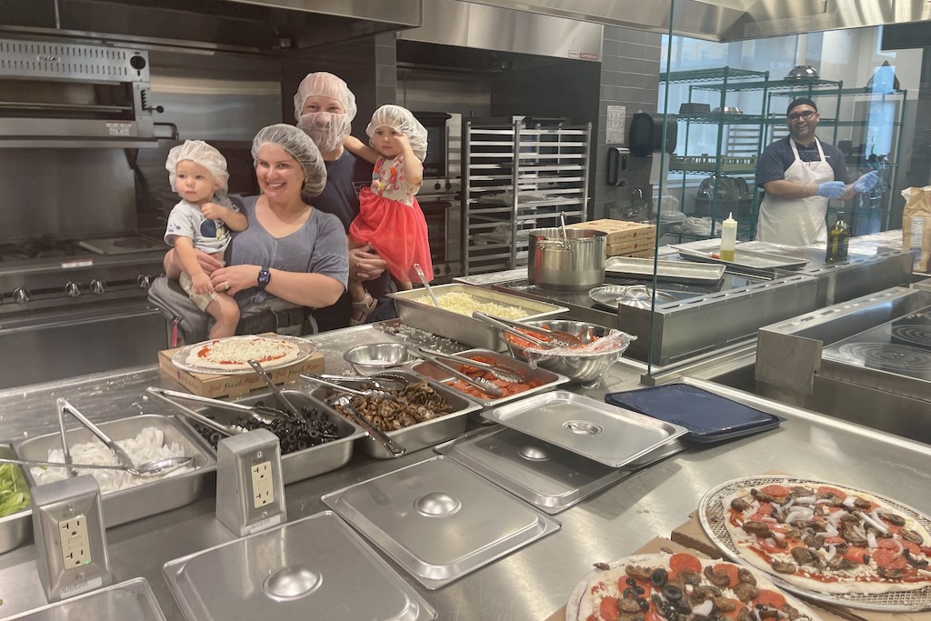 People in hairnets pose in front of prepped pizzas in an industrial kitchen.