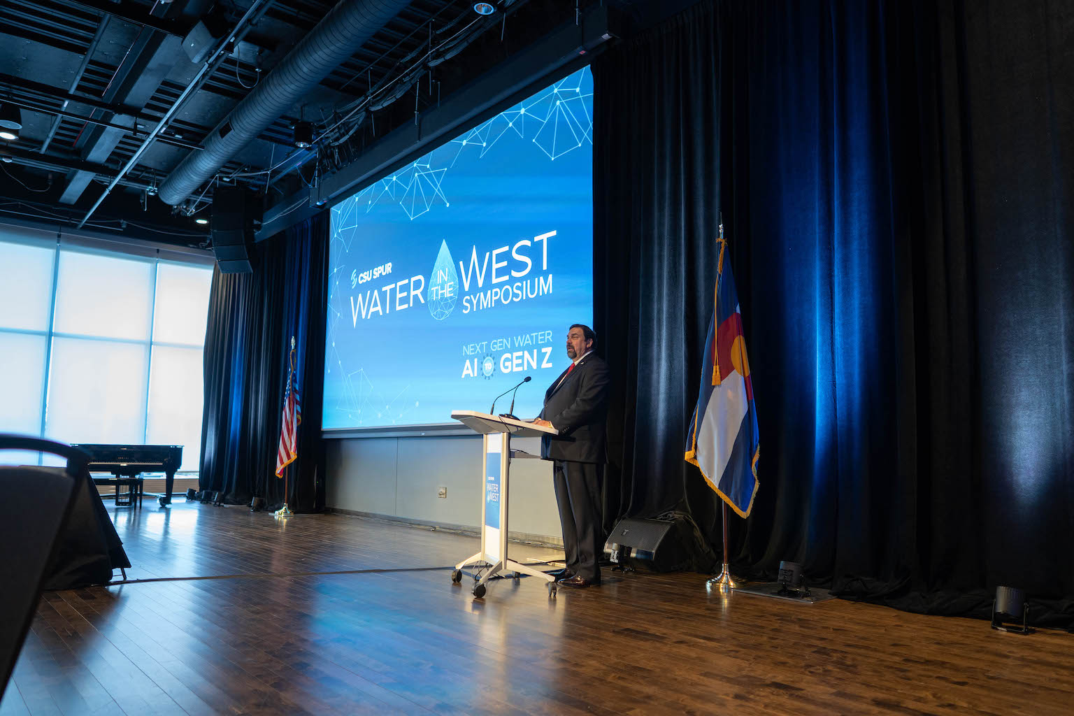 A man stands at a podium near a large screen with the Water in the West Symposium logo.