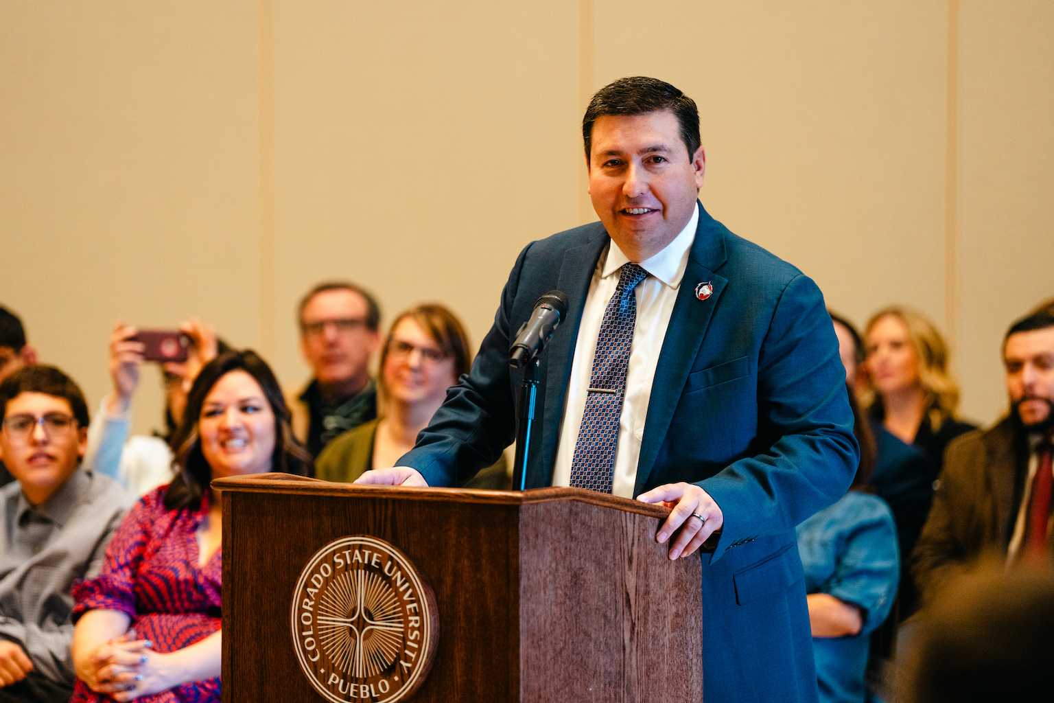 A man in a blue suit with a CSU Pueblo lapel pin stands at a podium.