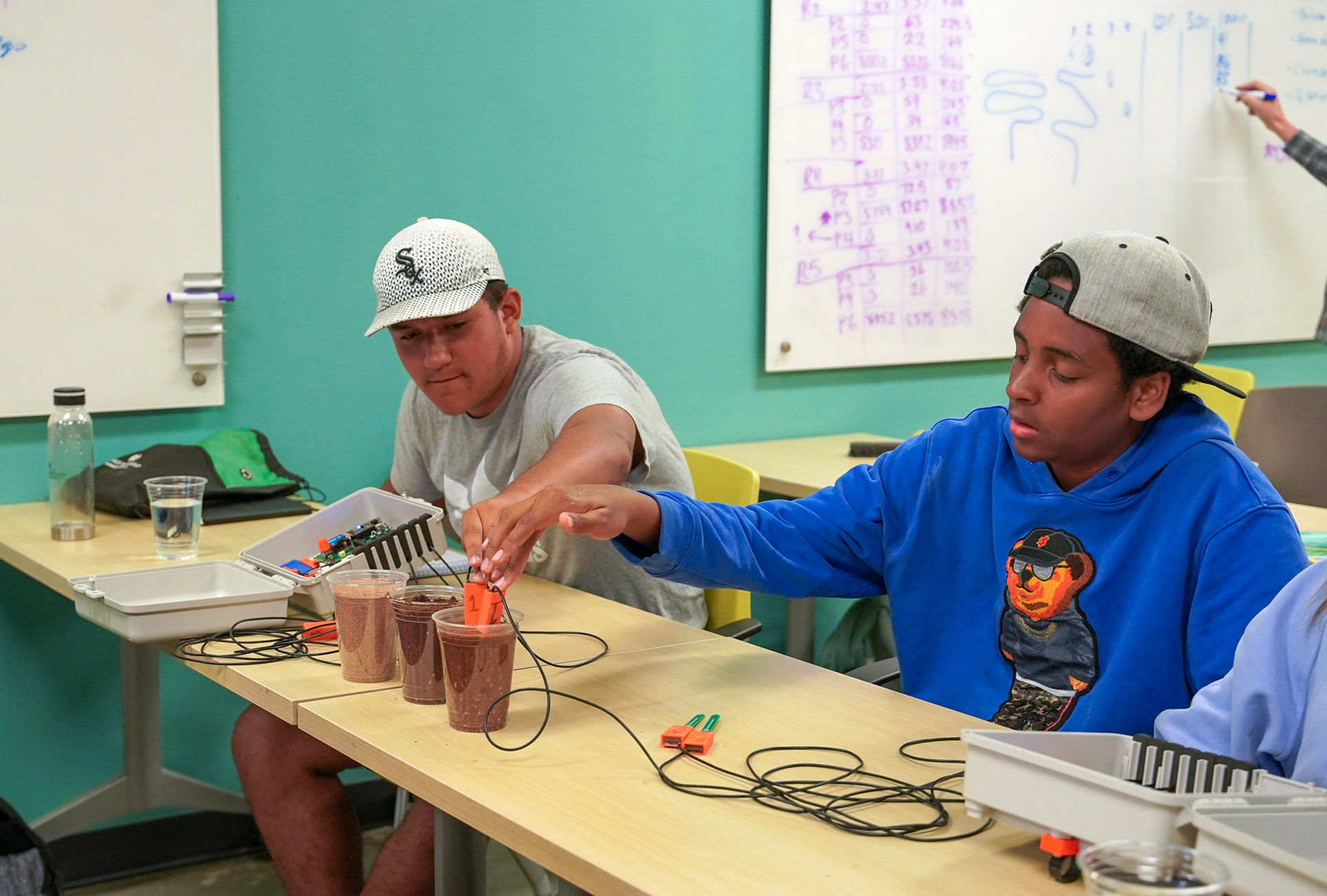 Two teenage boys in ball caps place devices connected with wires into soil samples in plastic cups in a classroom.