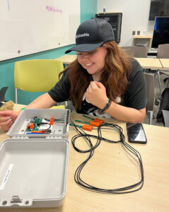 A smiling young woman looks inside a plastic box that contains technological equipment with wires running from inside the box to sensors on small soil stakes outside the box.