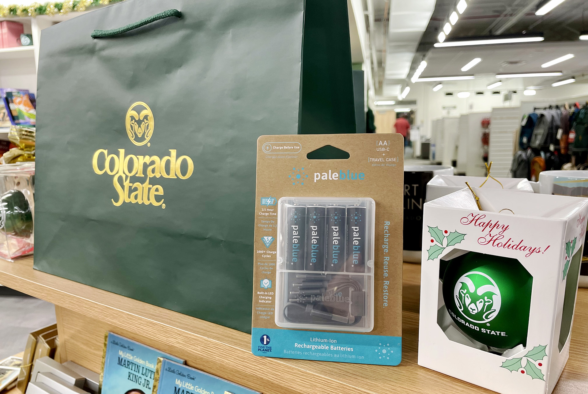 A four-pack of Paleblue rechargeable batteries sits between a CSU bag and CSU tree ornament. The batteries are on sale at CSU's Bookstore.