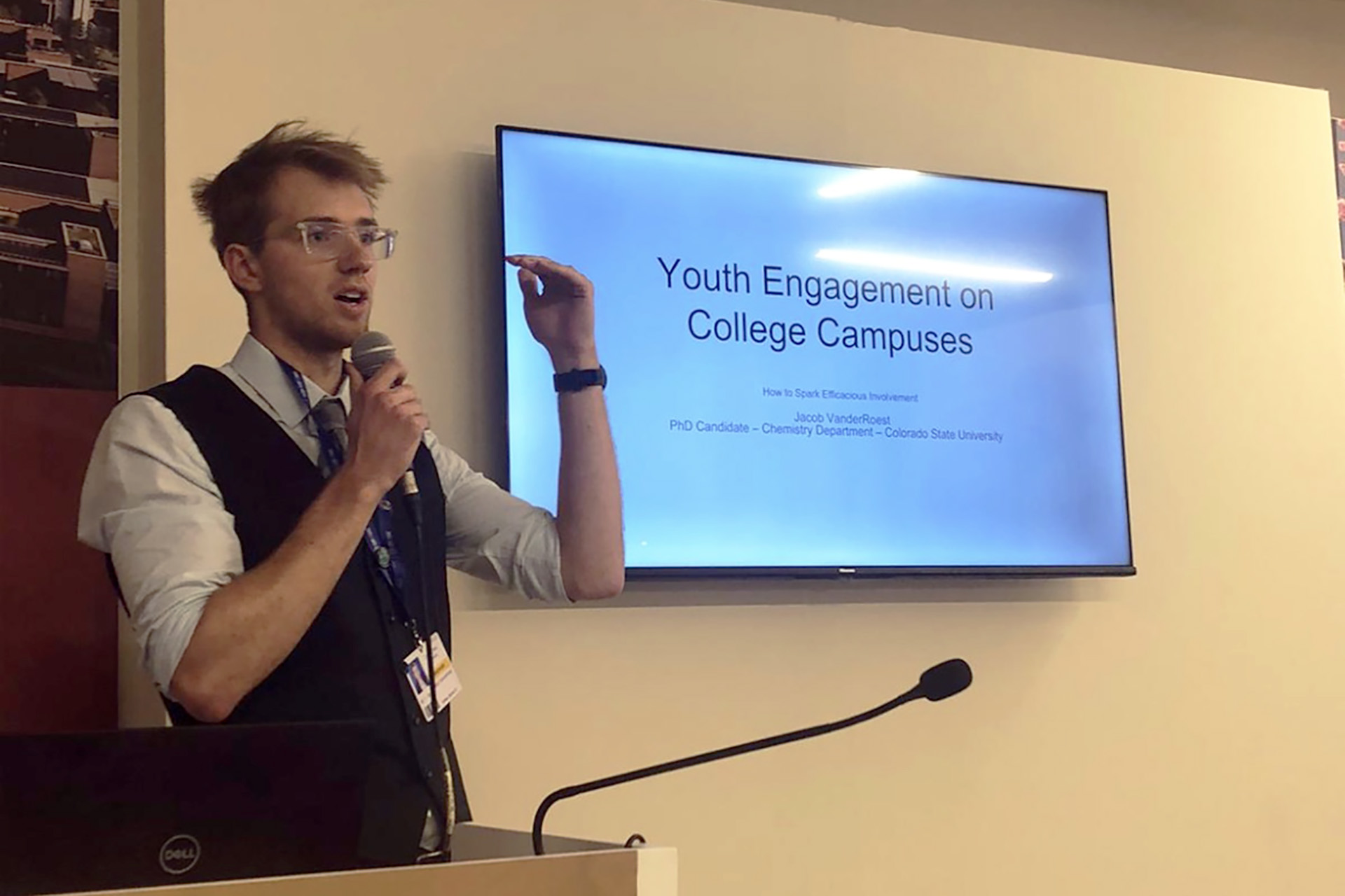 CSU student Jacob VanderRoest gives a presentation during COP28 in Dubai. The photo shows him speaking at a podium with a TV screen over his left shoulder.