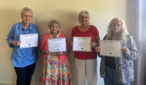 Four elderly women stand in a row, holding certificates and smiling for the camera.